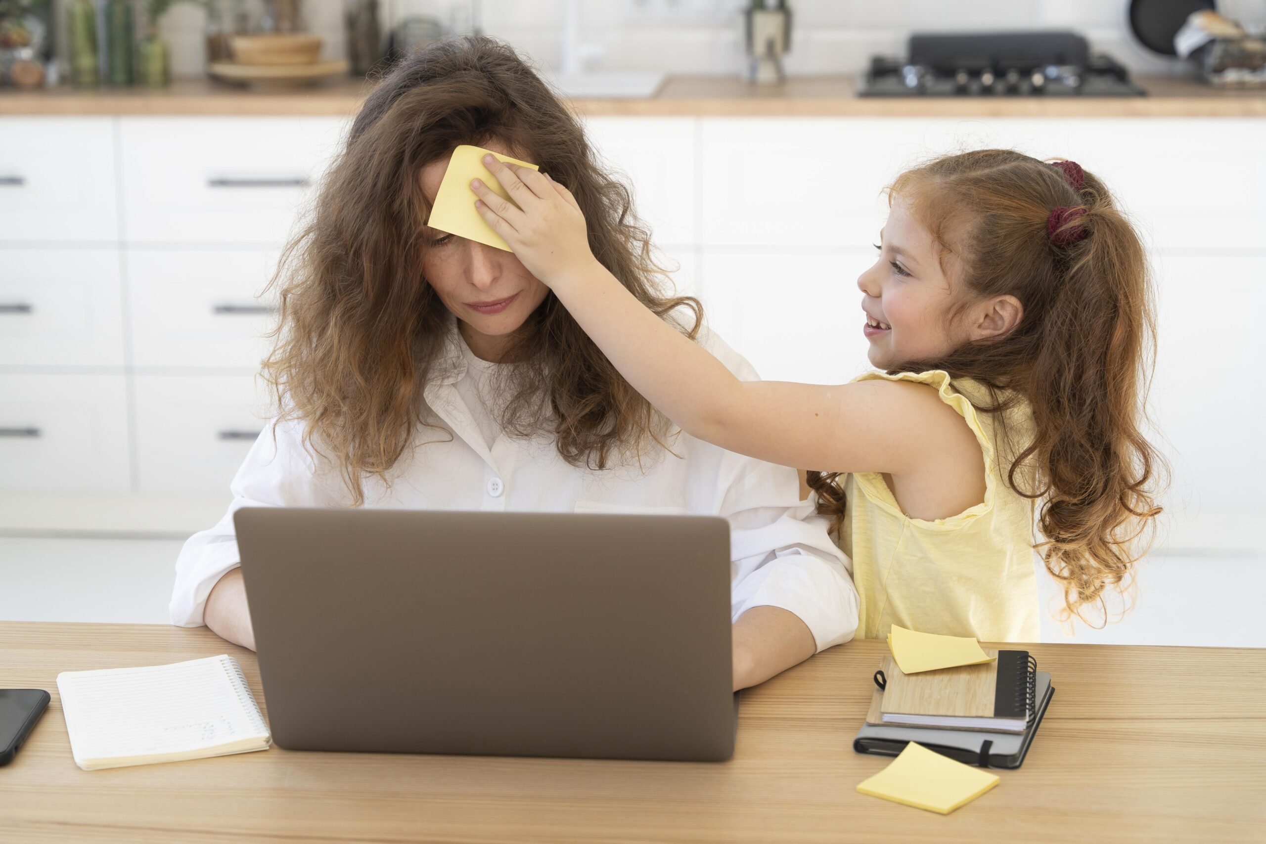Mujer mirando el computador preocupada mientras una niña pequeña le seca el sudor de la frente